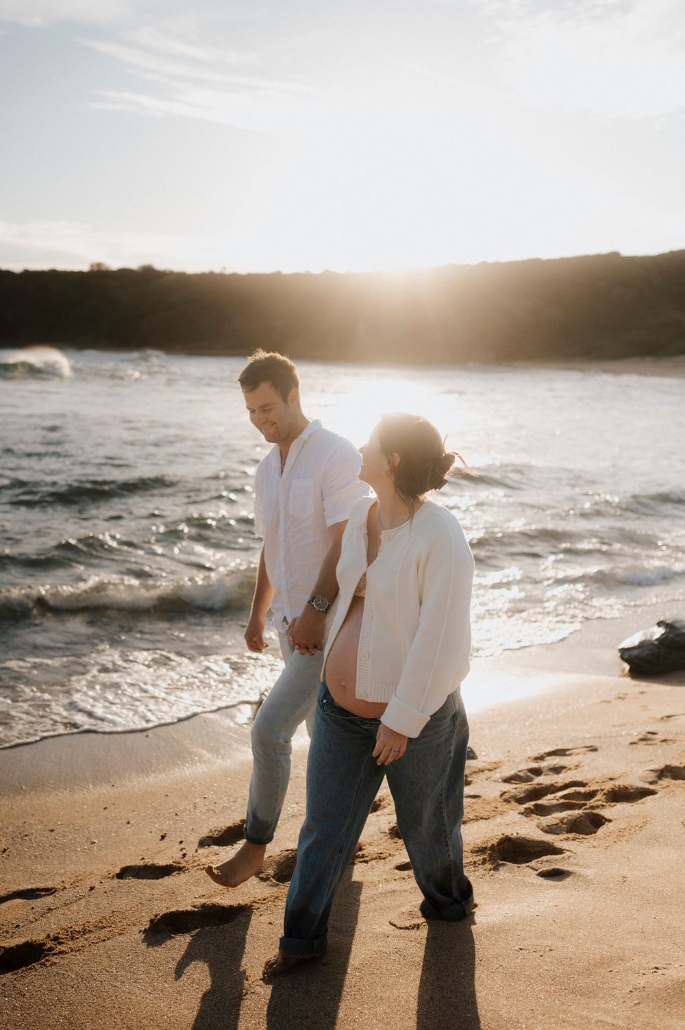 A couple walks along a sandy beach at sunset with ocean waves and cliffs in the background.