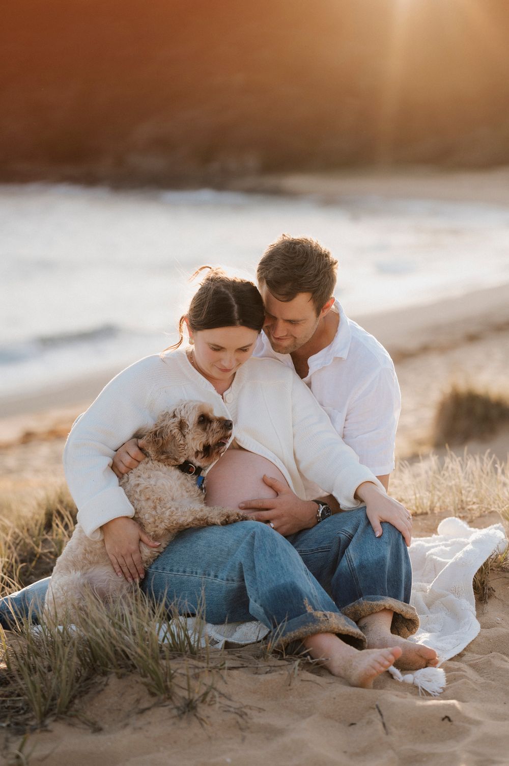 A couple sits on a sandy beach at sunset with their small dog while showing affection to a pregnant belly.