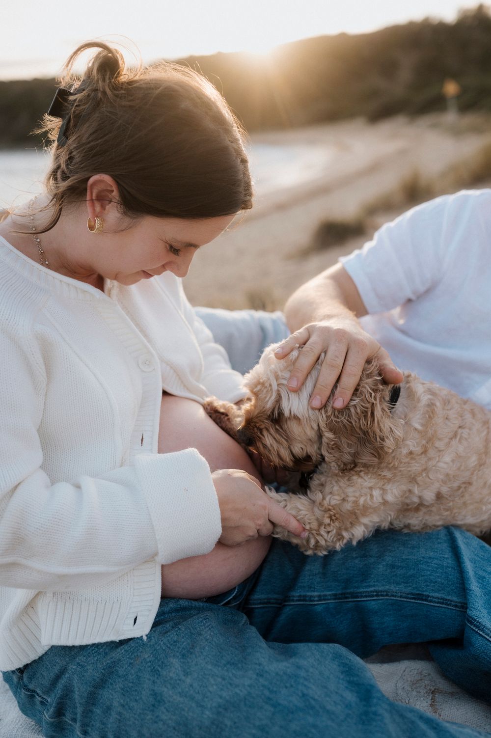 Person in a white sweater sharing a tender moment with a fluffy brown dog at sunset on the beach.