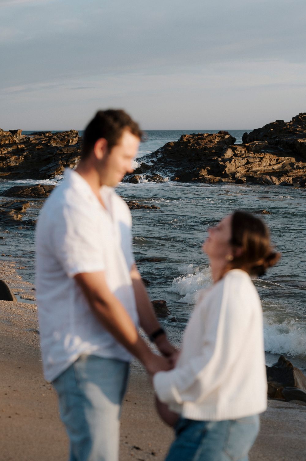 A couple in white shirts share a romantic moment on a rocky beach at sunset with ocean waves crashing behind them.