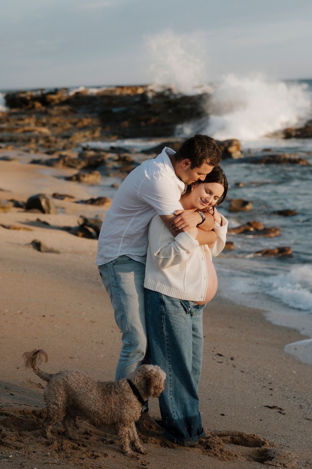 A romantic couple embraces on a rocky beach with crashing waves and ocean spray in the background.