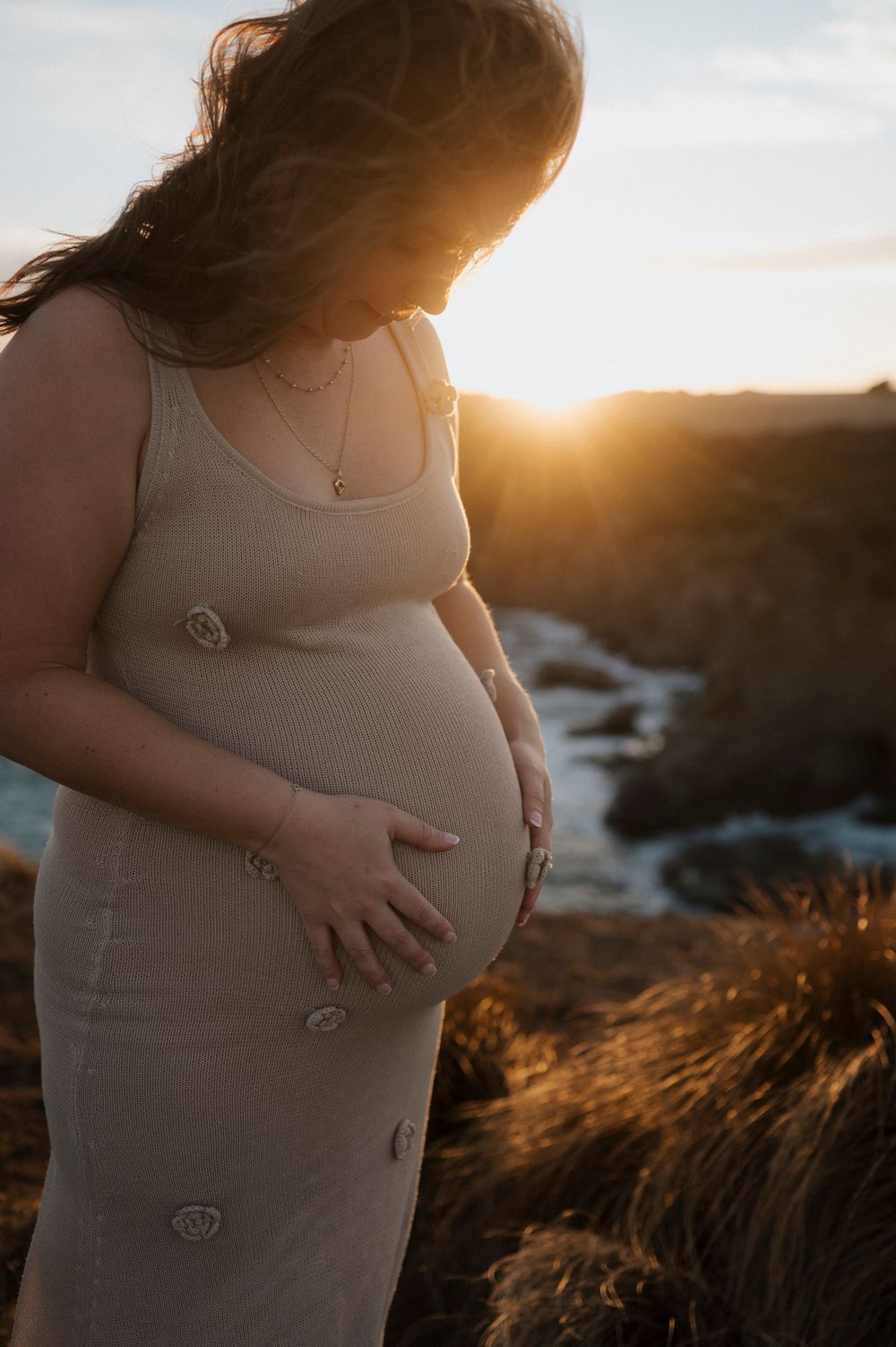 A pregnant person in a white dress caresses their belly during golden hour sunset, silhouetted against nature.