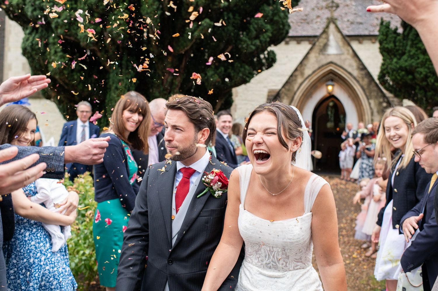 Couple exit a church as petal confetti is thrown, the bride is laughing