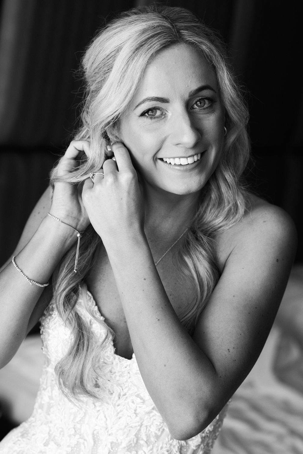 A black and white portrait of a smiling person with wavy blonde hair wearing a white lace dress and jewelry.