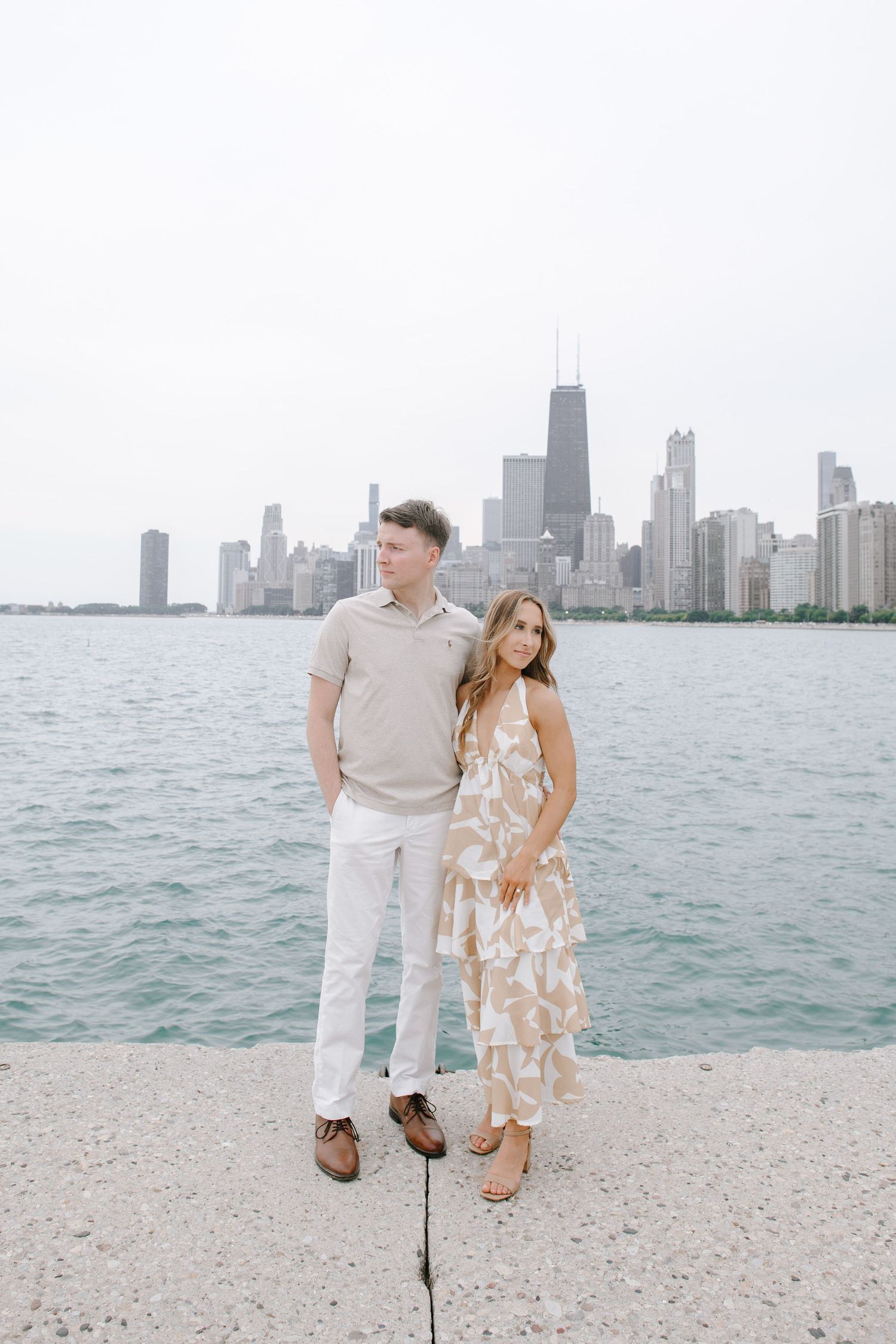 A couple stands together on a beach with the Chicago city skyline visible across Lake Michigan.