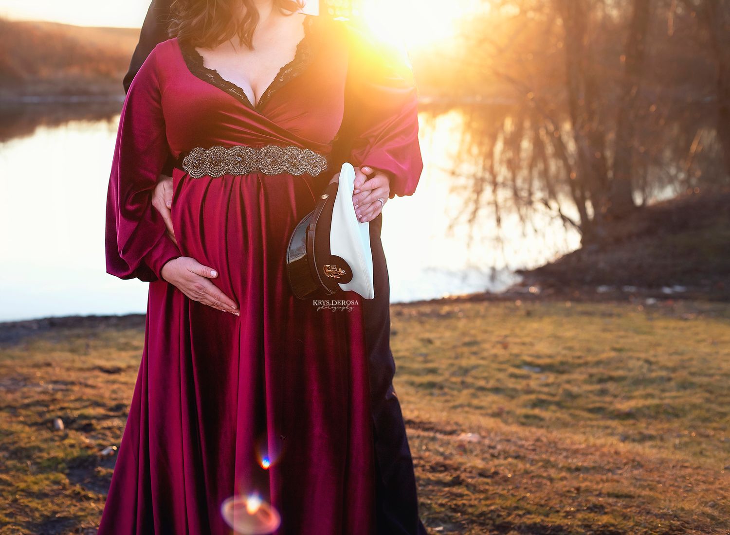 Pregnant woman in flowing burgundy dress stands by lake at sunset holding wine glass.
