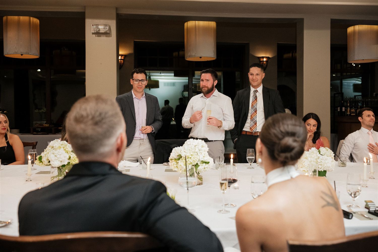 A formal wedding reception with guests seated at tables adorned with white linens and lit cylindrical pendant lights.