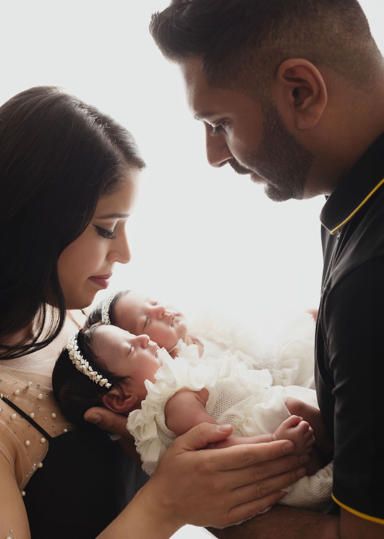Elizabeth Lang Photography newborn photos mom and dad hold newborn twin girls in a bright backlit photo with little white dresses.