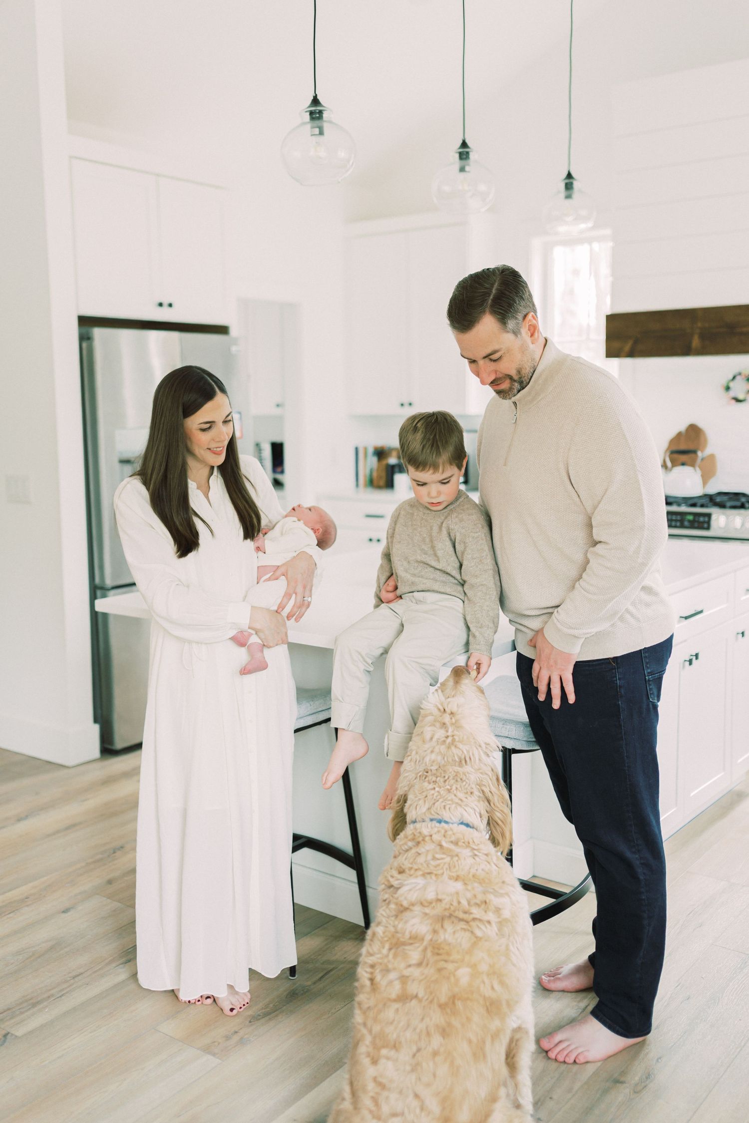 Family gathered in modern white kitchen with large goldendoodle dog on hardwood floors and pendant lighting above.