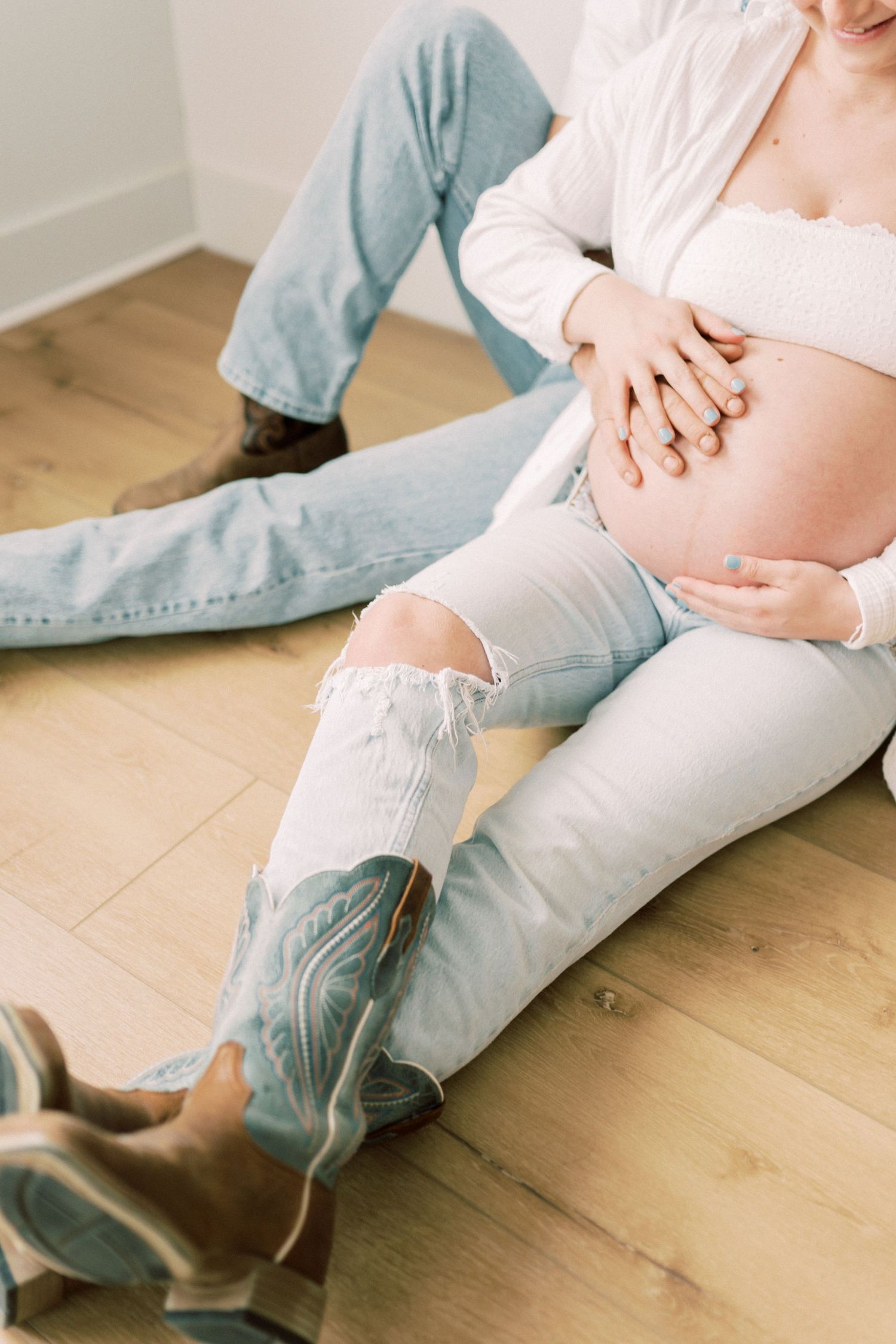 Pregnant person in ripped jeans sits on hardwood floor wearing cowboy boots while cradling their baby bump.