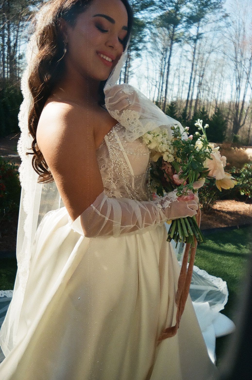 A bride in a flowing ivory gown with sheer lace sleeves holds a delicate bouquet of white flowers in an outdoor setting.