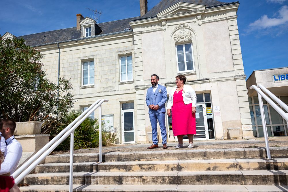 A couple stands on stone steps outside a grand white building wearing bright blue and pink outfits.