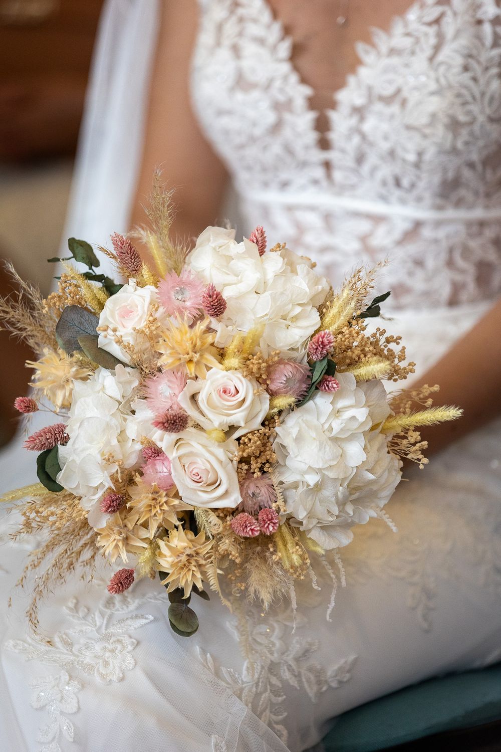 Elegant bridal bouquet featuring blush roses, white hydrangeas, and dried flowers resting on lace wedding dress.