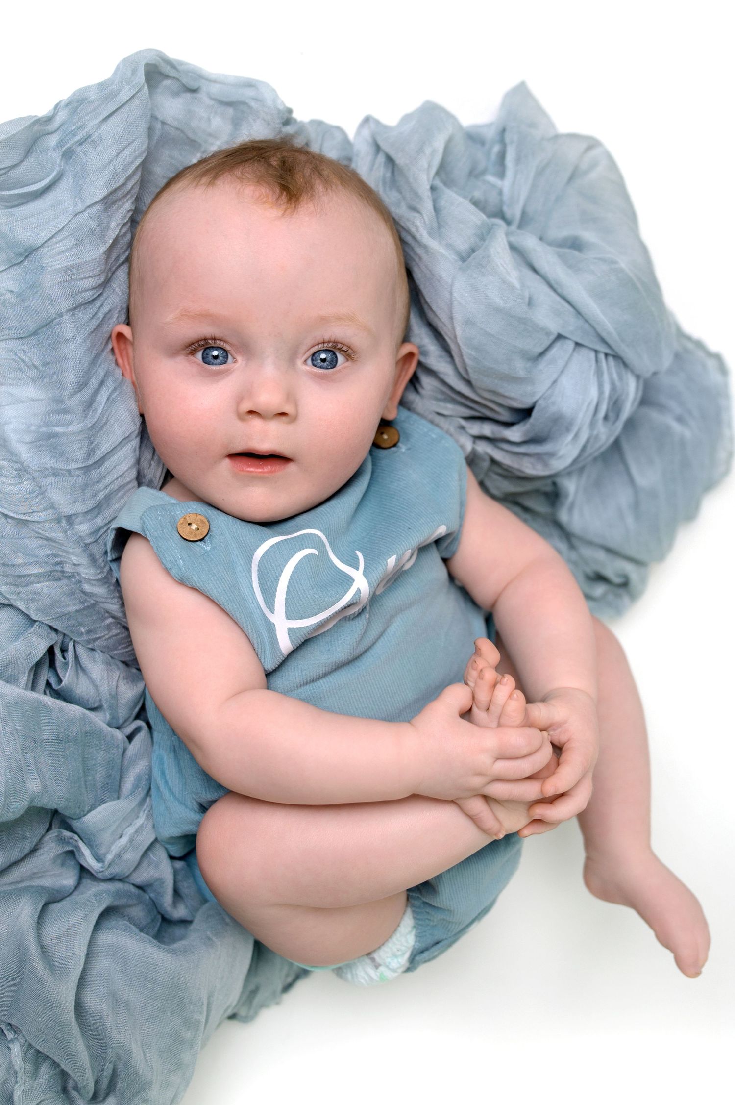 Baby in light blue romper lying on soft blue fabric with bright eyes and clasped hands.