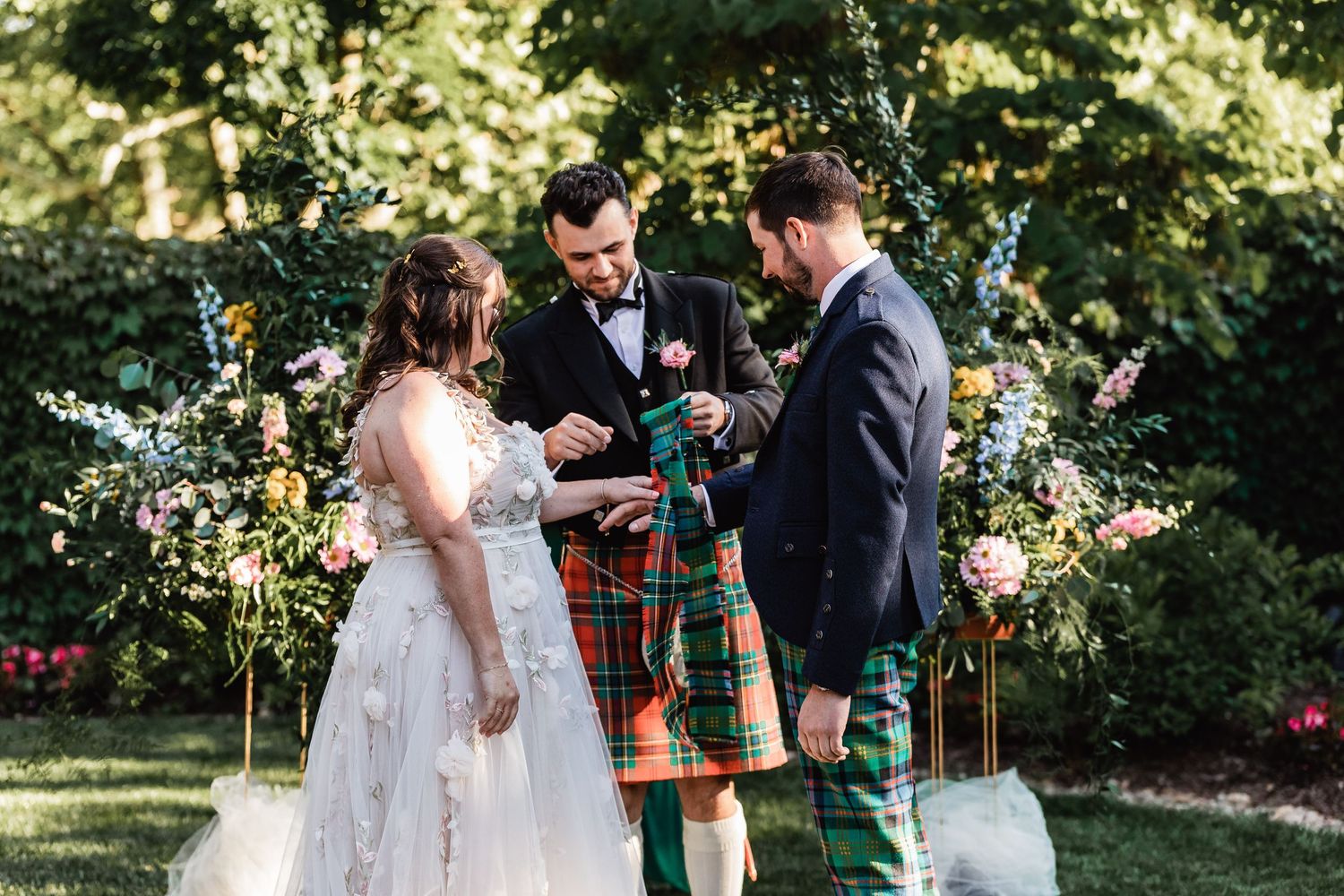 Scottish wedding ceremony takes place in a beautiful garden with traditional kilts and lace wedding dress on sunny day.