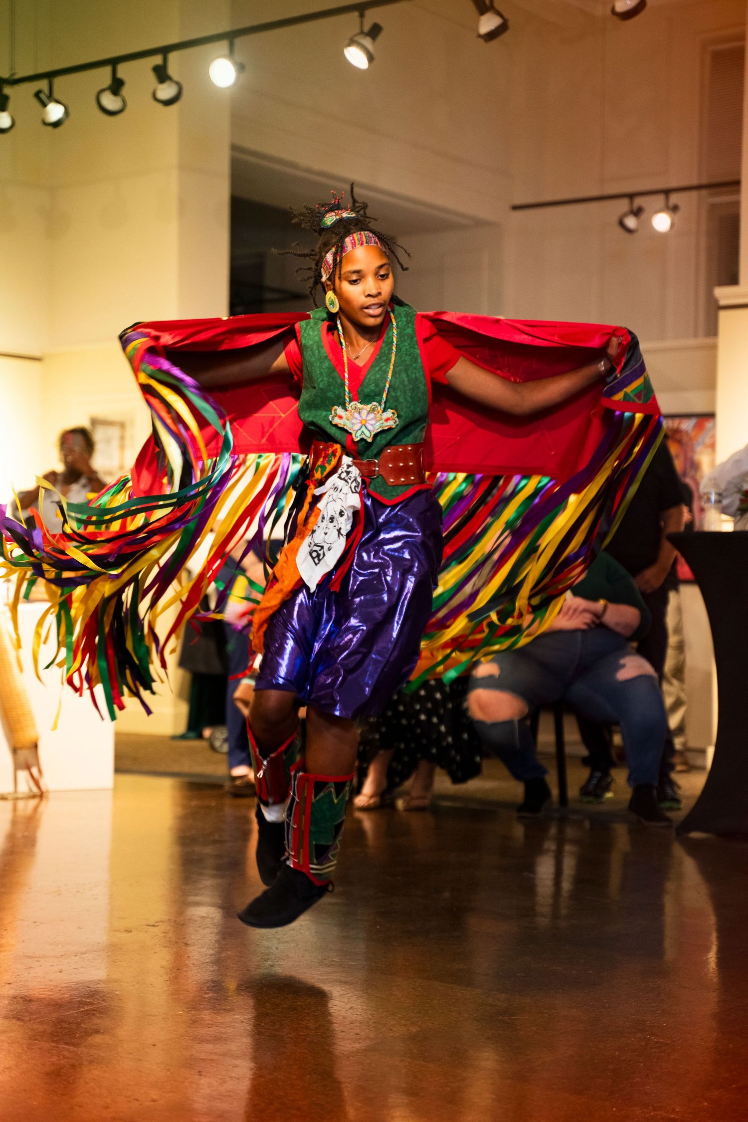 Dancers perform in colorful flowing costumes on a gallery floor under spotlights during an artistic performance.