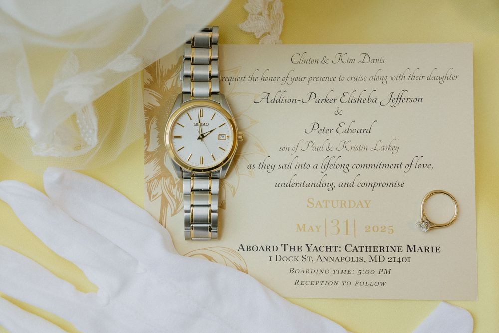 A silver wristwatch sits atop an elegant wedding invitation with gold accents and delicate floral details in the background.