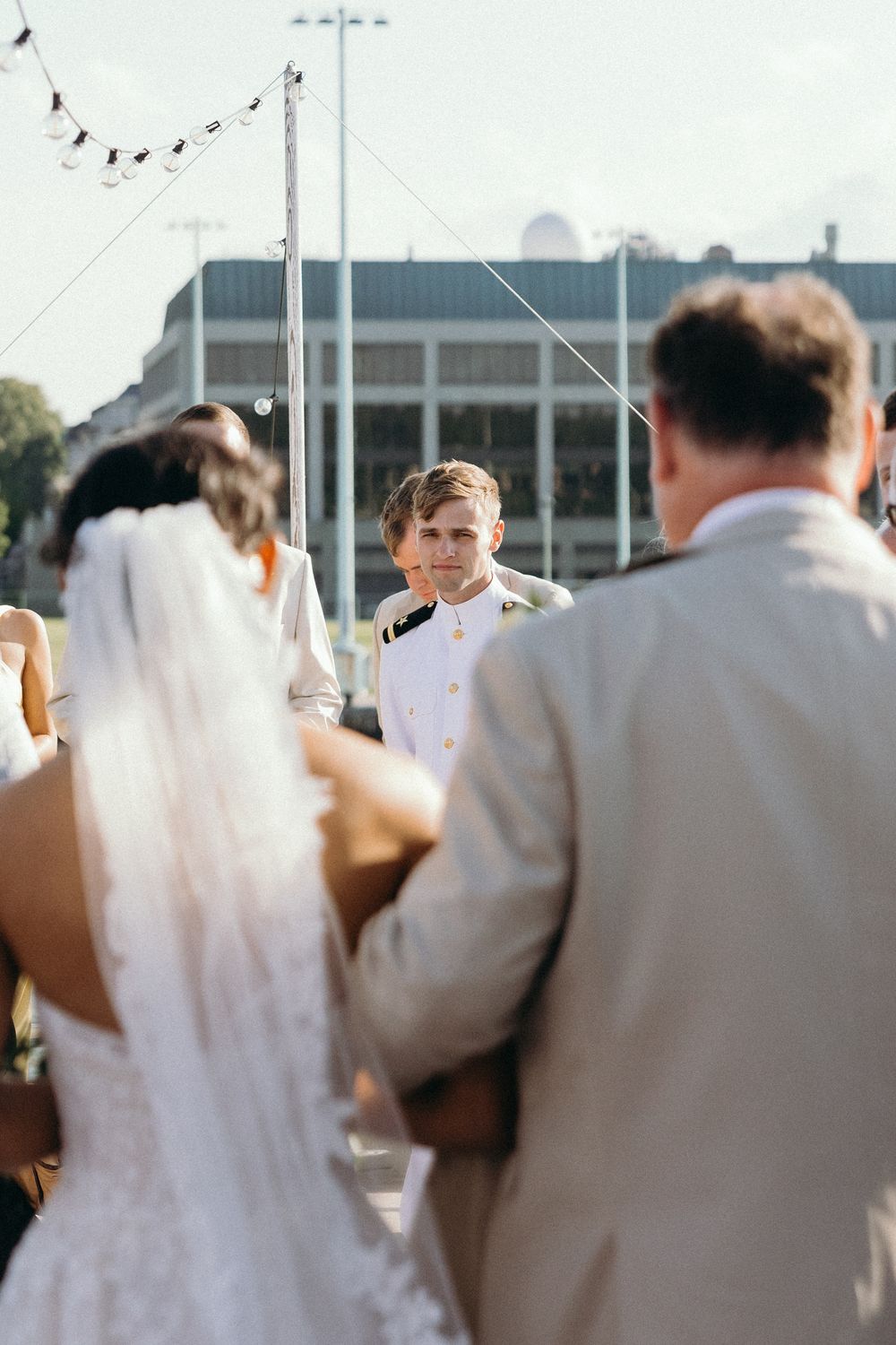 An outdoor wedding ceremony takes place with attendees gathered under bright sunlight near a modern building.