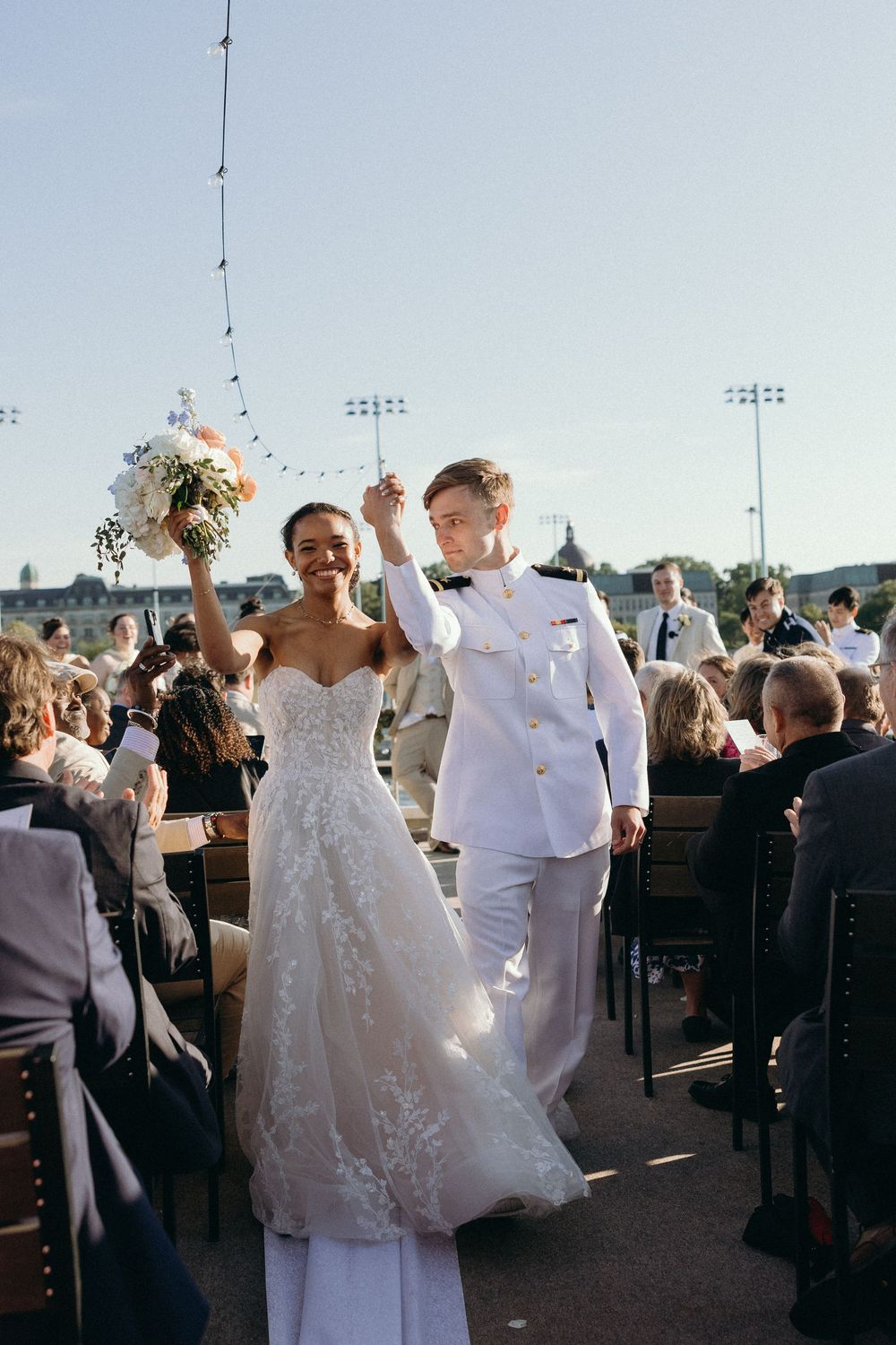 A newlywed couple walks down the aisle during an outdoor ceremony, surrounded by guests and stadium floodlights.