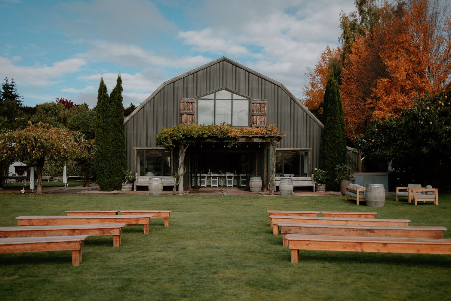 A rustic barn wedding venue with wooden benches arranged on a green lawn for an outdoor ceremony.
