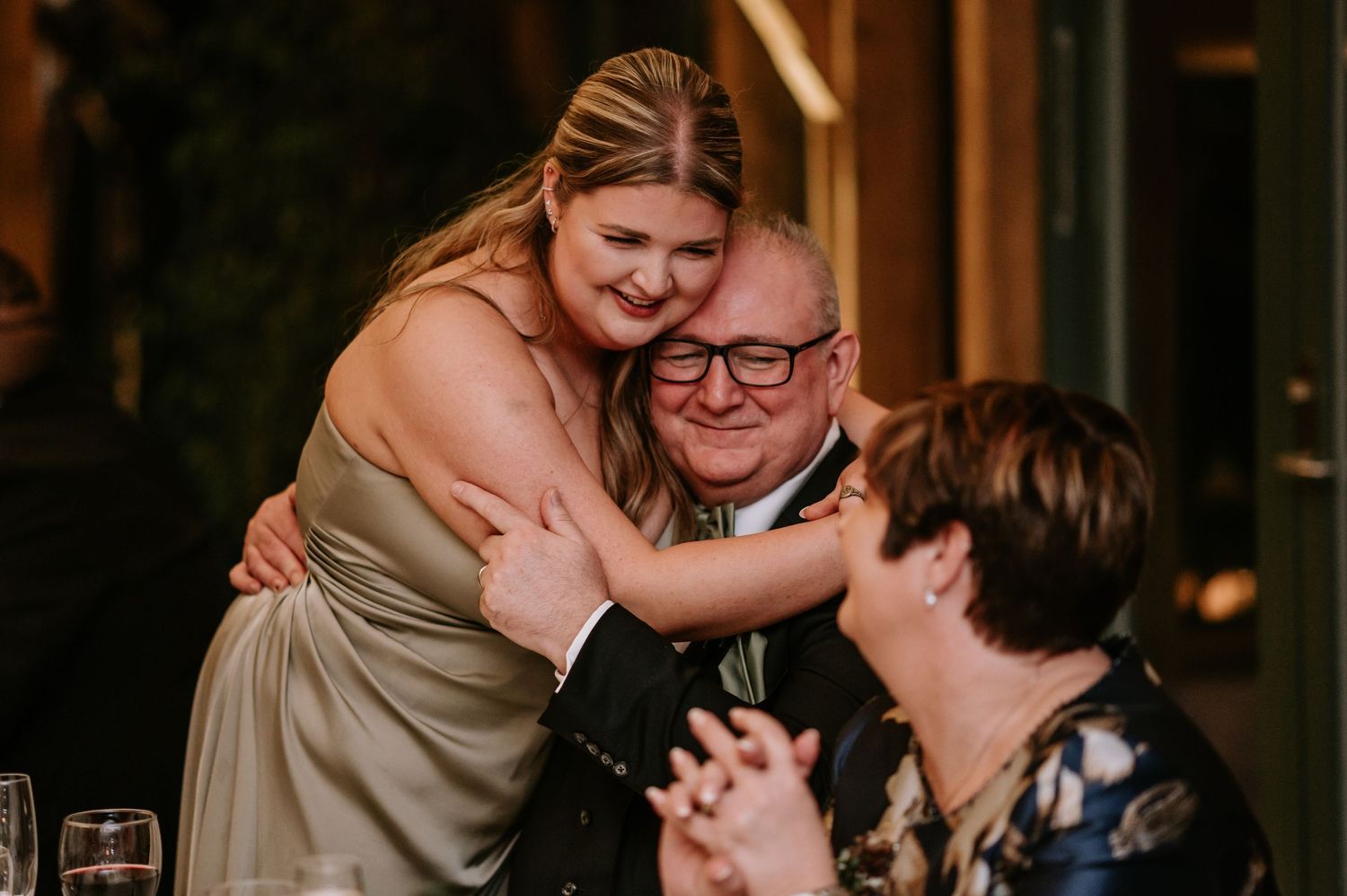 Heartfelt moment of a bridesmaid hugging her father on the wedding day, with him smiling at his wife.