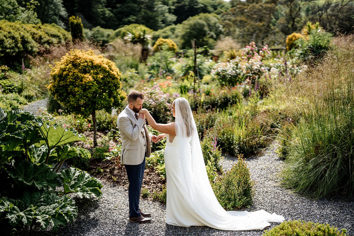A couple stands together on a winding garden path surrounded by lush greenery and blooming flowers in a sunny outdoor setting.