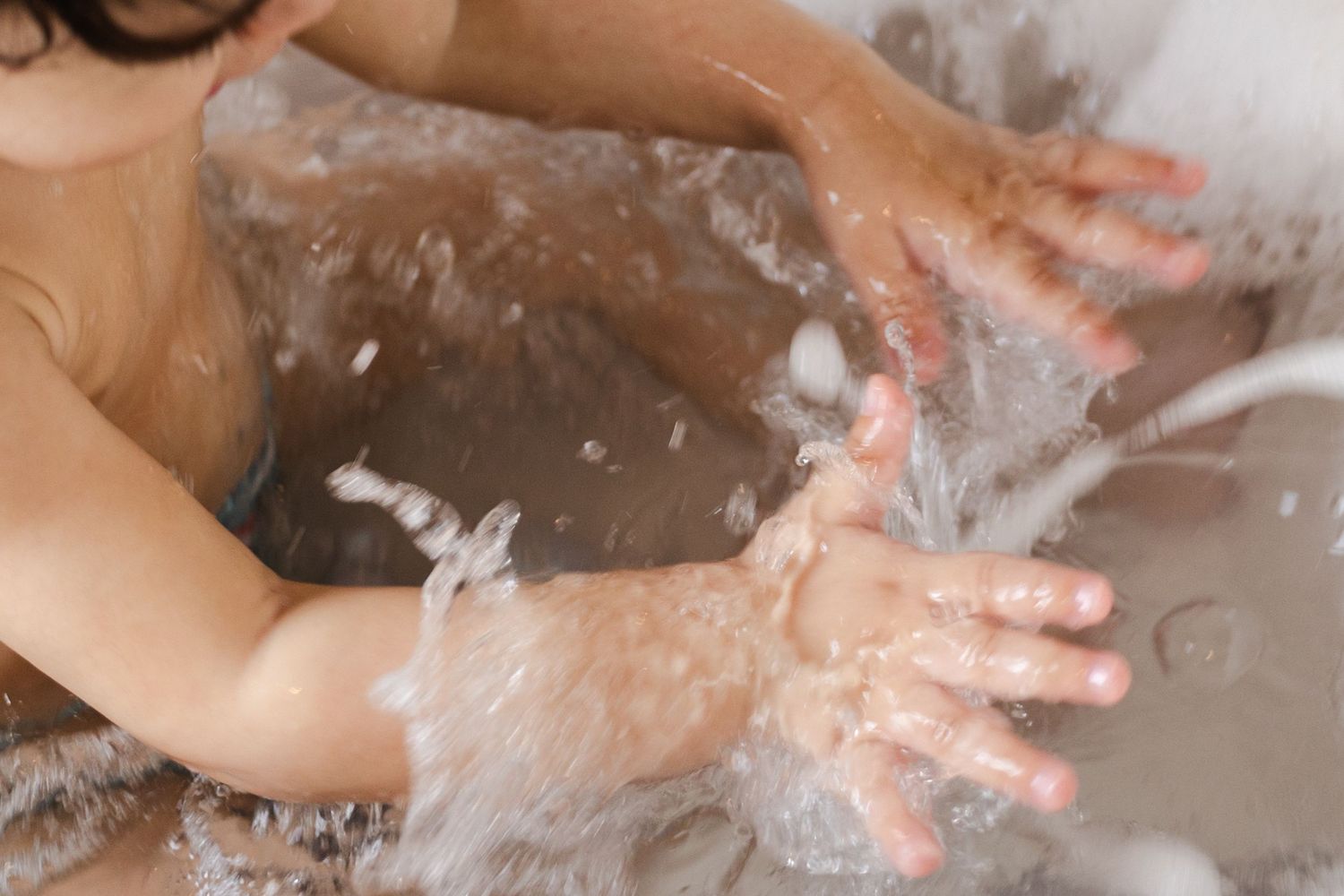 Small hands splash and play with soap bubbles and water in a white porcelain sink.