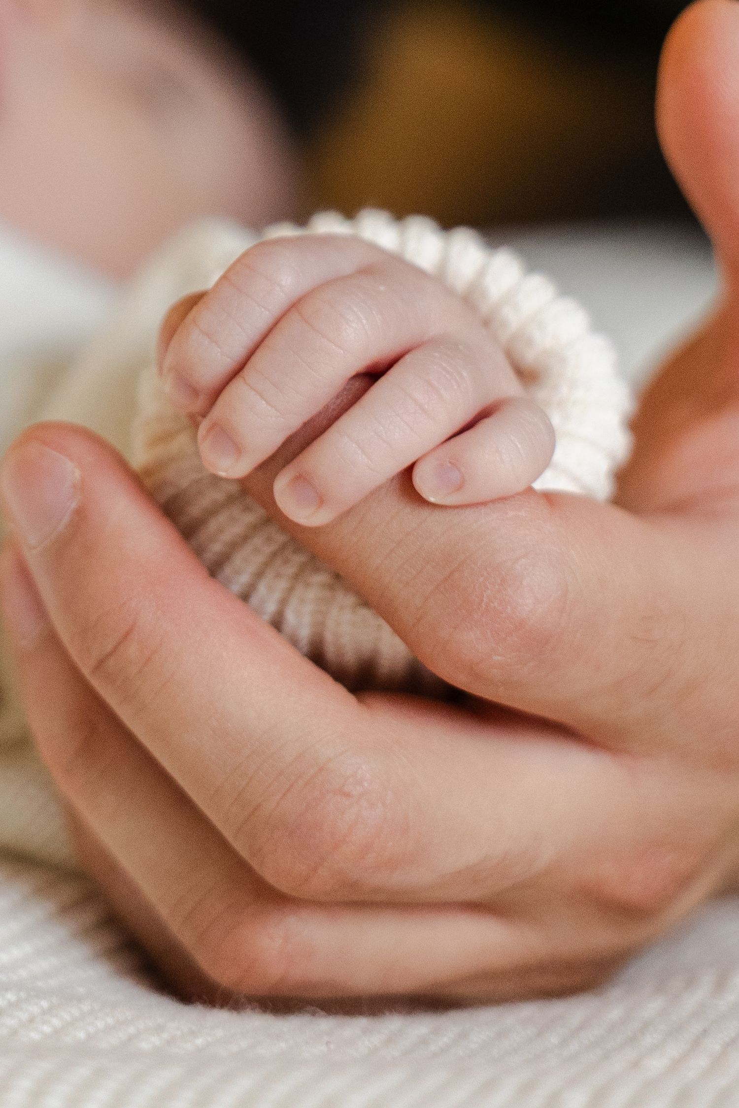 A tiny baby's hand gently rests in an adult's palm against a soft white knit fabric background.