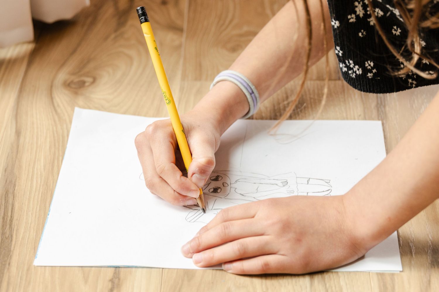 Student writing or drawing on white paper with a yellow pencil on wooden desk surface.