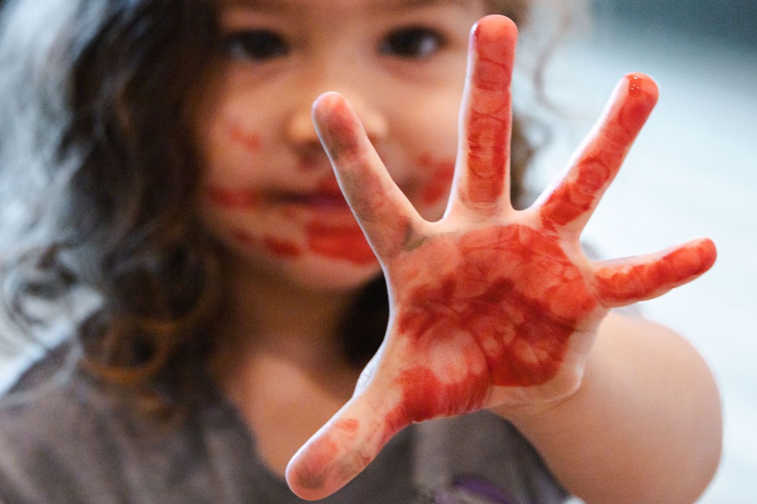 Child shows palm of hand covered in bright red paint against blurred background.