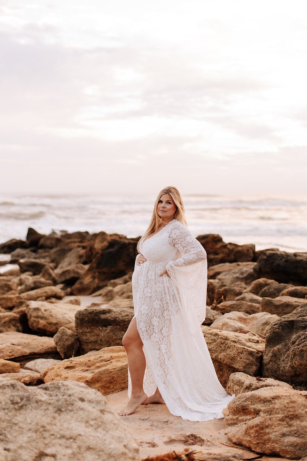 Woman in flowing white lace dress poses on rocky beach during sunset.