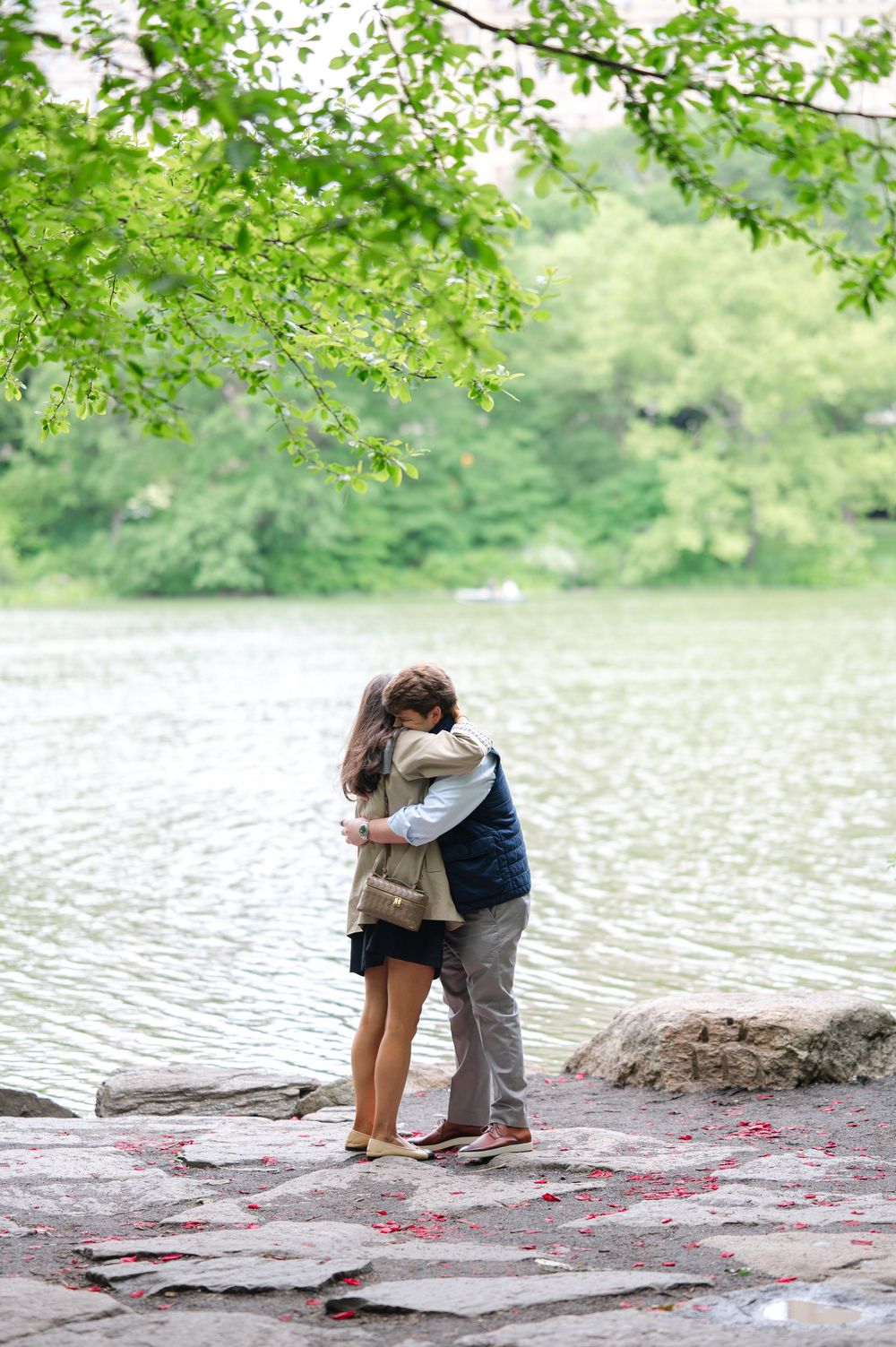 Two people share a romantic embrace on rocky outcrop by serene lake while green tree branches frame the scene from above.