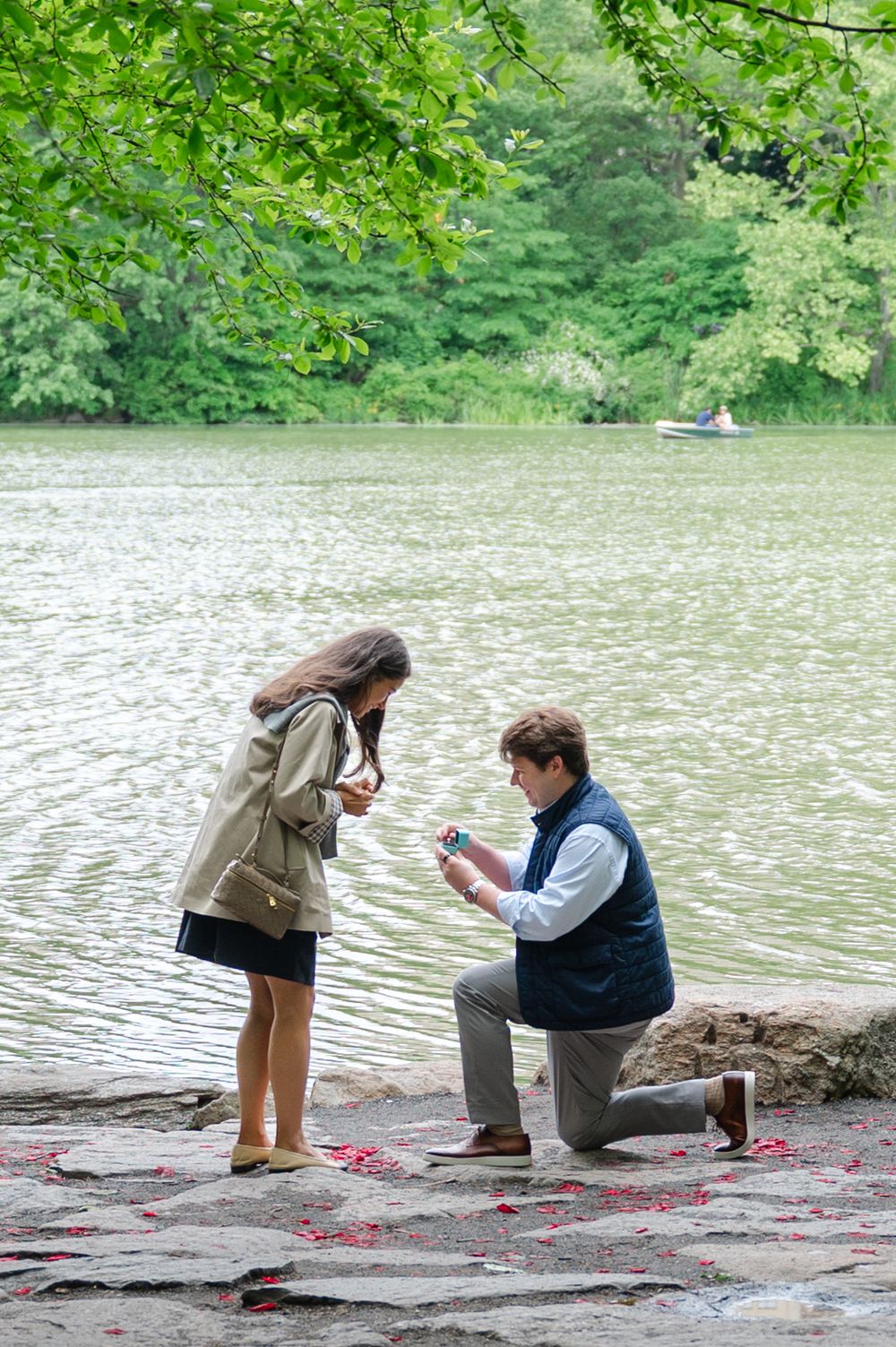 A romantic proposal scene by a calm lake surrounded by lush green trees on a peaceful summer day in Central Park