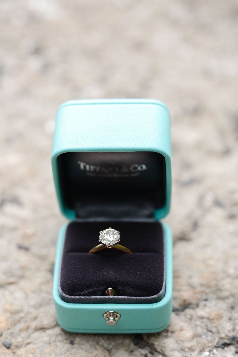 A sparkling diamond engagement ring sits in an iconic Tiffany & Co. turquoise blue box on a stone surface.