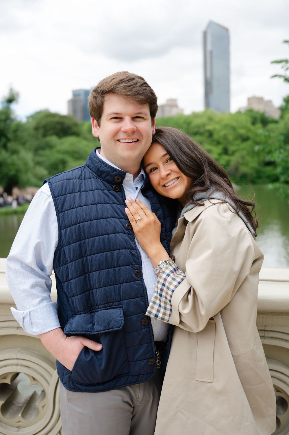 Happy couple embraces on a stone bridge in Central Park with Manhattan skyline in background during NYC proposal