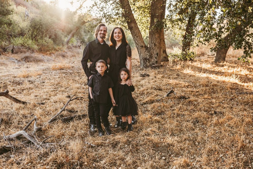 A loving family in black outfits poses together on a sunny hillside with trees in the background.