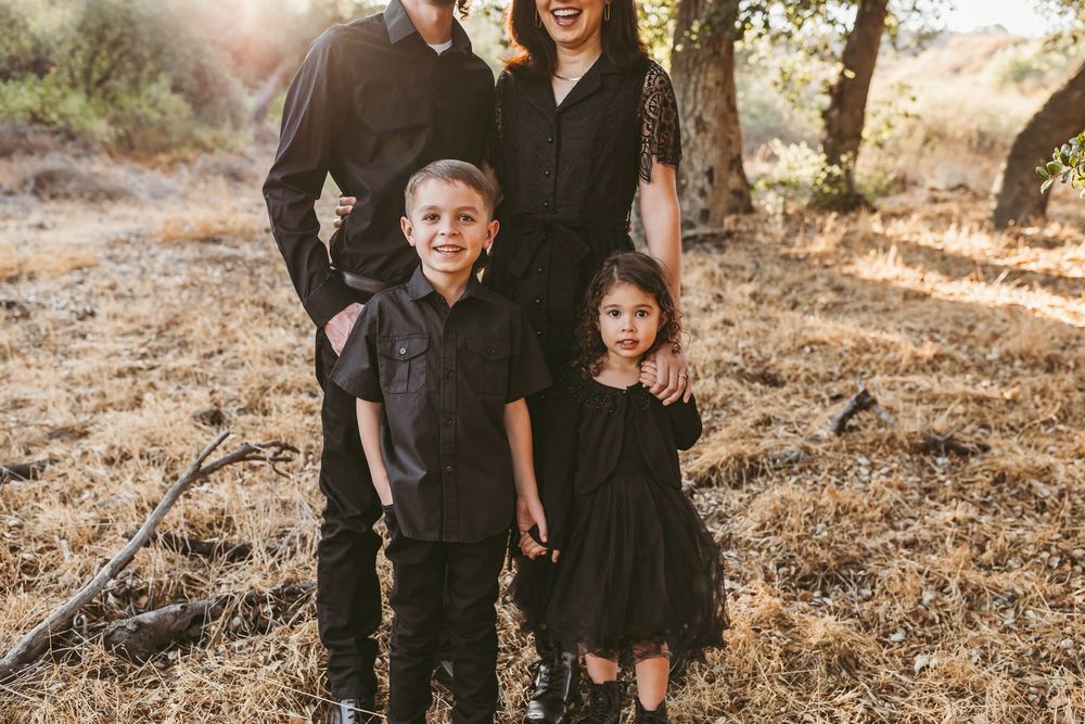 A family of four dressed in coordinating black outfits poses outdoors in a natural setting during golden hour.