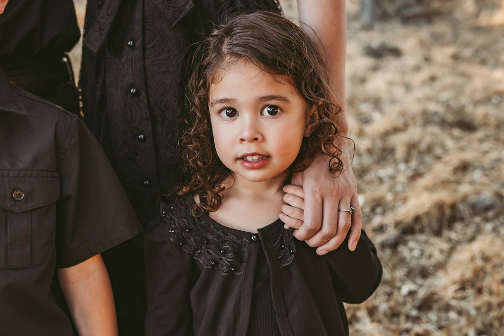 Two children wearing black outfits pose together in a rustic outdoor setting with dry grass and natural lighting.