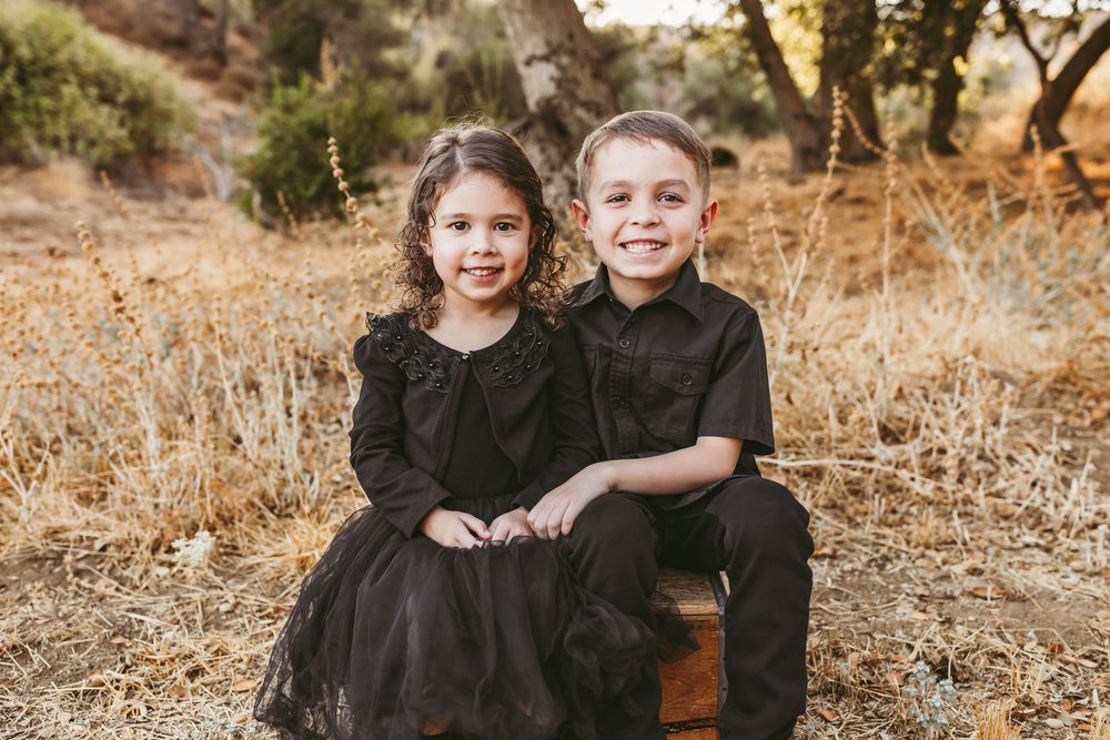 Siblings in matching black outfits sit together outdoors against a natural autumn landscape background.
