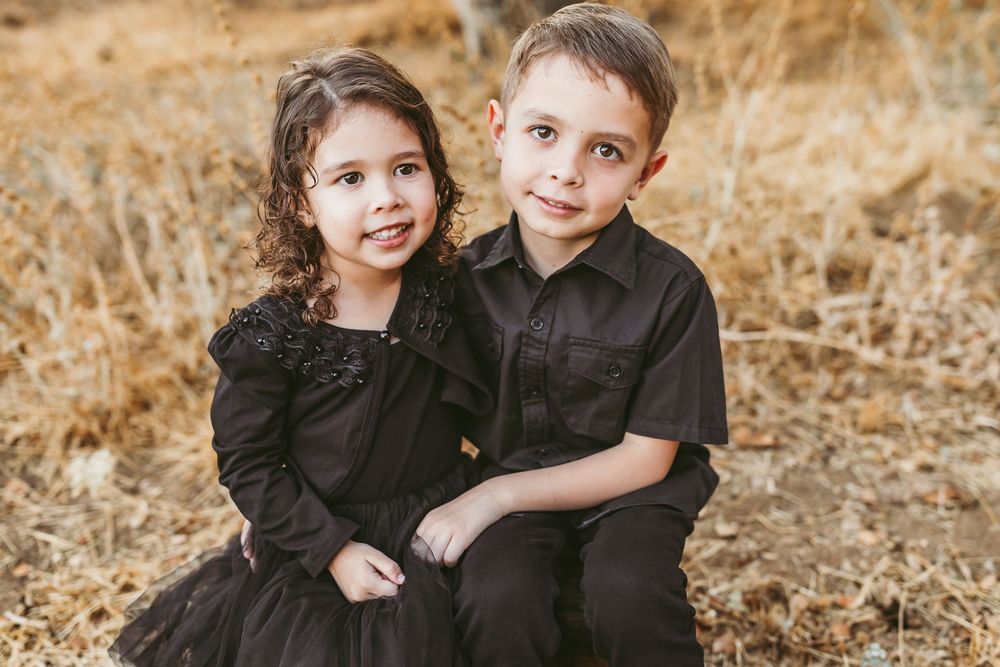 Two young children dressed in dark clothing share a candid moment while sitting in a field of golden grass.