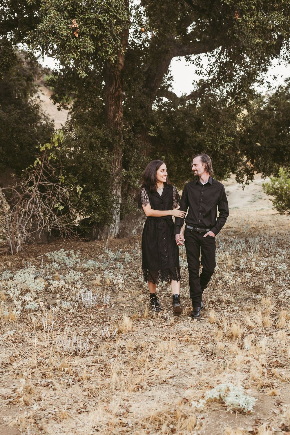 A couple in matching black outfits walking together through a serene outdoor setting with oak trees.