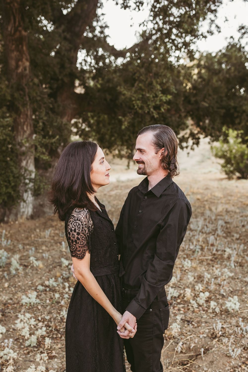 A couple dressed in black holds hands and gazes at each other lovingly while standing in a scenic outdoor setting.