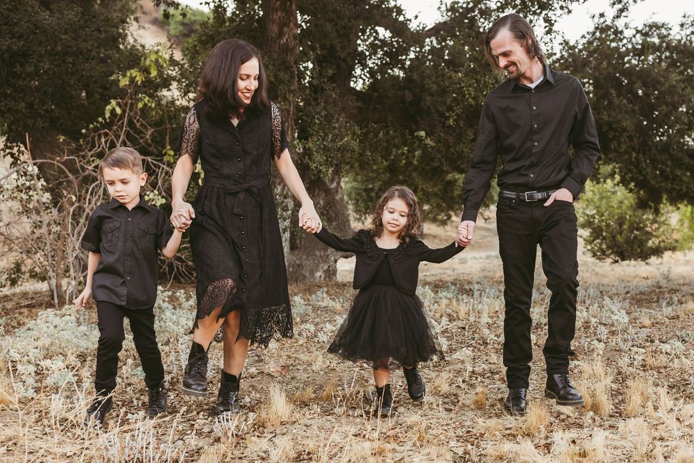 Family in coordinated dark outfits holds hands while standing together in outdoor natural setting.