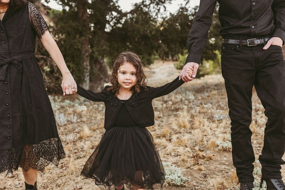 A young girl in a black dress holds hands with her parents while standing in a rustic outdoor setting.
