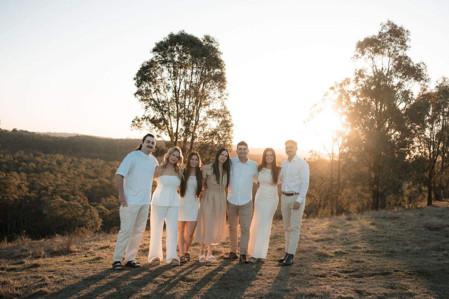 A group stands together outdoors at sunset wearing coordinated white and beige outfits against a backdrop of eucalyptus trees.