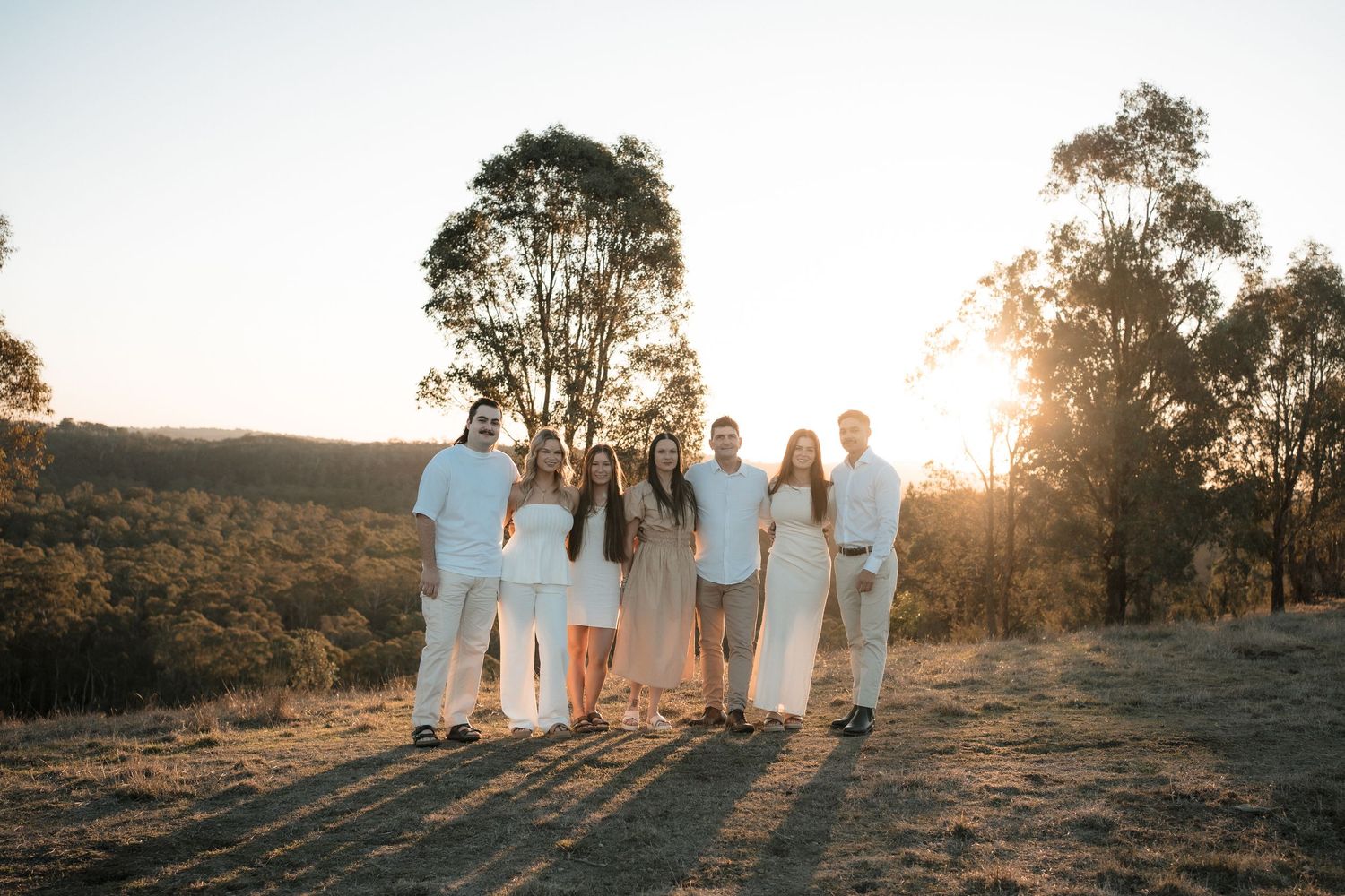 A group in white outfits stands together on a hilltop at sunset while silhouetted eucalyptus trees frame the scene.