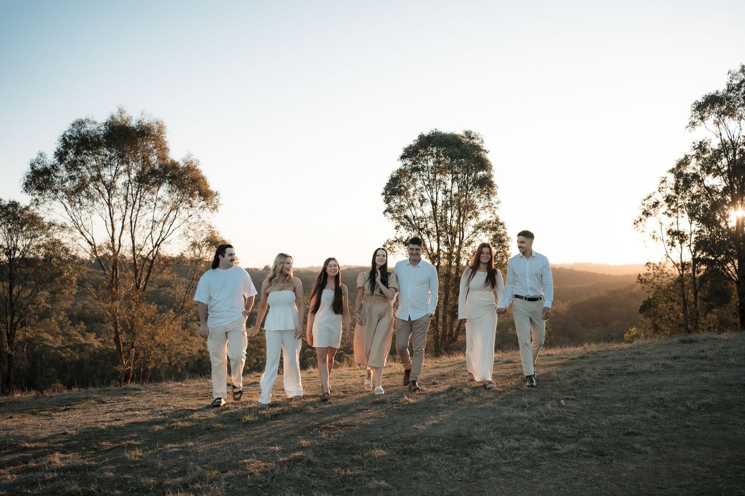 A group wearing white outfits stands together on a hilltop at sunset with eucalyptus trees in the background.