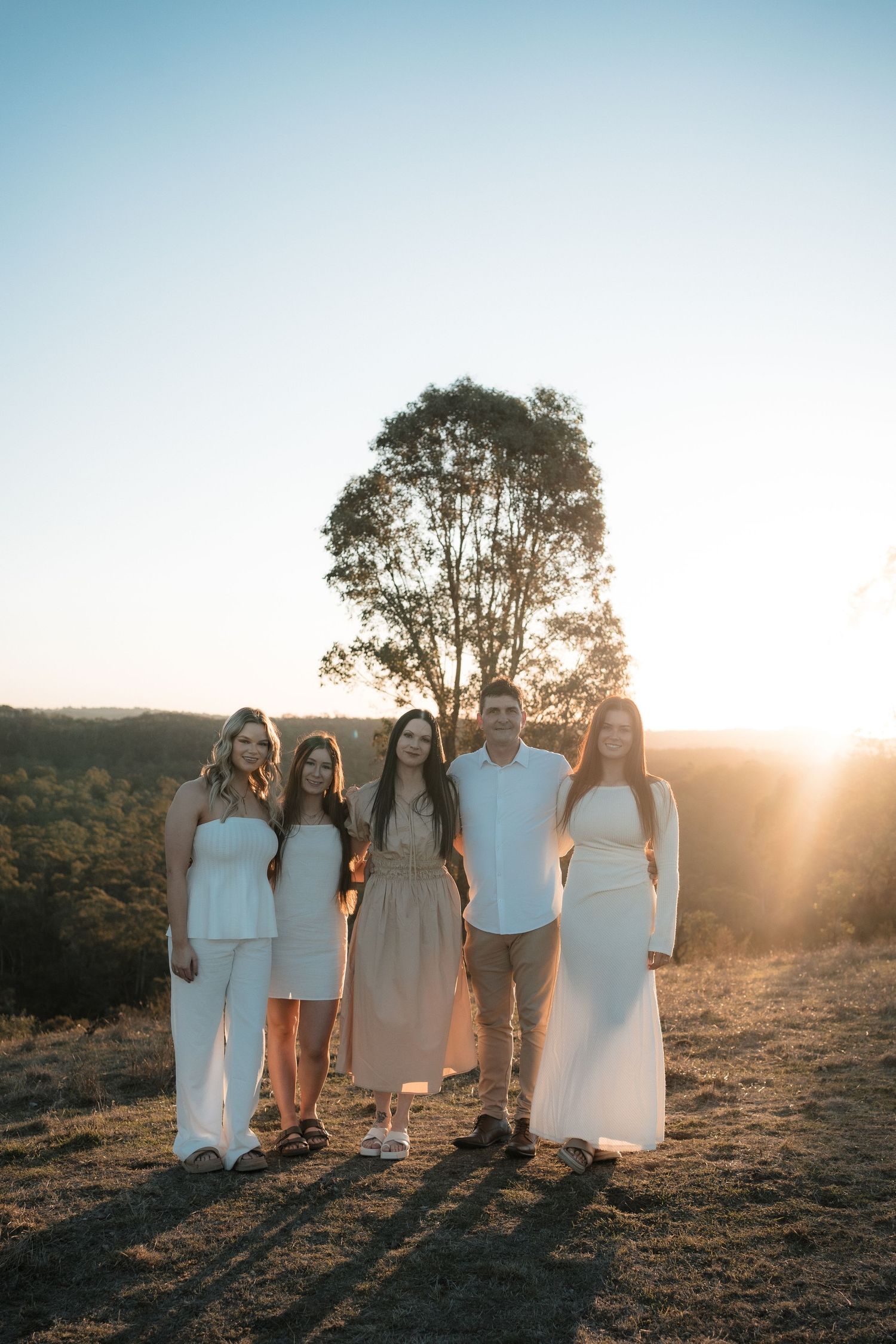 A group of people in white outfits pose together on a hilltop during golden hour sunset with a tall tree behind them.