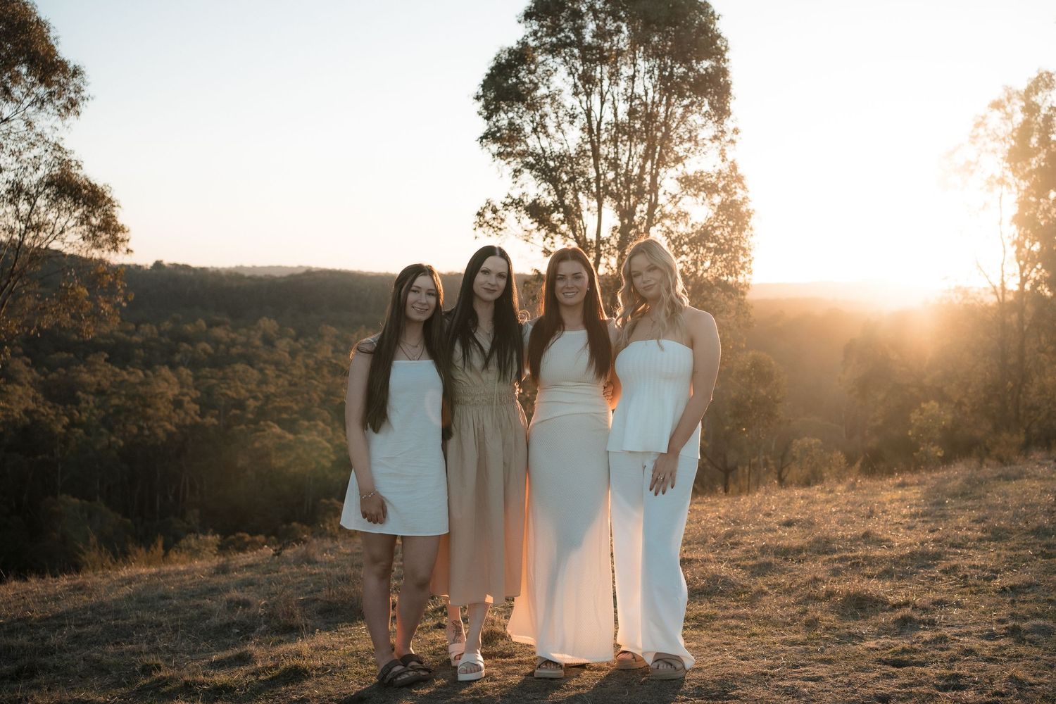 Four people in white dresses pose together at sunset in a natural outdoor setting with trees and rolling hills.