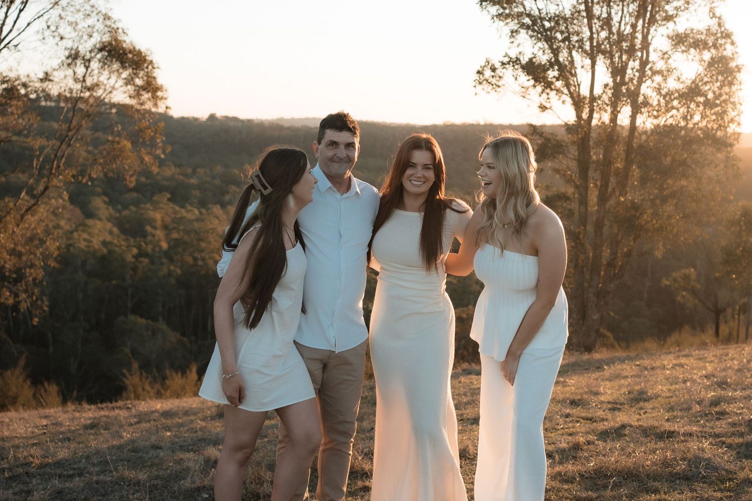 Group in white attire standing together on a hillside during golden sunset with trees in background.